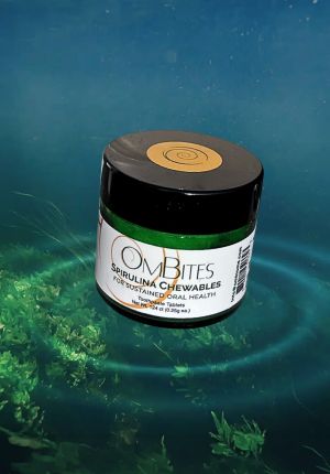 A small jar of OmBites Spirulina Chewables toothpaste tablets floating above underwater green algae in blue water.