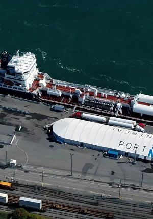 Aerial view of a tanker berthed alongside a white domed warehouse, storage tanks, tank trucks, and rail lines.