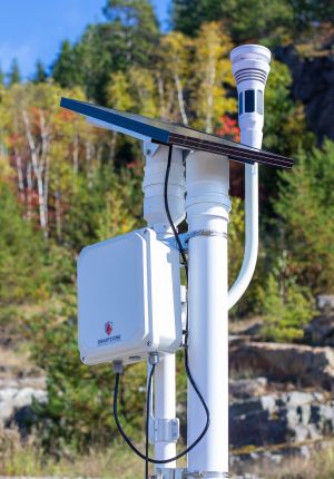 A white SmartCone sensor enclosure mounted on a pole with a solar panel and weather sensor, installed outdoors against autumn forest.