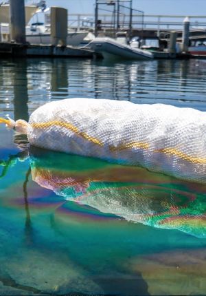 A white mesh sorbent boom floating in marina water, absorbing an iridescent rainbow-colored oil sheen, with boats docked behind.