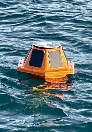 Orange trapezoidal buoy device with solar panels floating on calm teal-colored water.