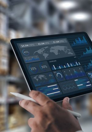 Hands holding a tablet displaying a dark analytics dashboard with revenue figures, world maps, donut charts, and trend graphs, in a blurred warehouse.